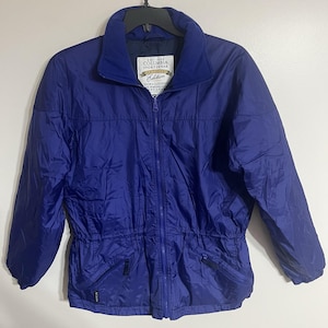 Puede incluir: Chaqueta azul Columbia Sportswear Edition con cremallera, dos bolsillos delanteros y cintura ajustable. La chaqueta tiene cuello alto y un pequeño parche con el logo. La chaqueta está colgada de una percha negra.
