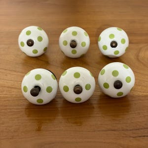 May include: Six white ceramic cabinet knobs with a green polka dot pattern. Each knob has a dark metal screw. The knobs are arranged on a wooden surface.