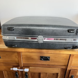 May include: A vintage black suitcase with silver-colored trim and metal clasps. The suitcase has a textured surface and rounded corners. It is sitting on a wooden dresser.