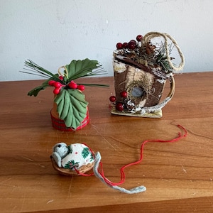 May include: Three holiday ornaments: a terracotta pot with green leaves and red berries, a rustic wooden birdhouse decorated with pine needles and red berries, and a small gray mouse figurine nestled in a walnut shell with a red string.
