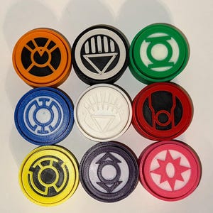 May include: Nine circular emblems in a variety of colours, including orange, black, white, green, blue, red, yellow, purple, and pink. Each emblem features a unique geometric design. The items appear to be 3D printed.