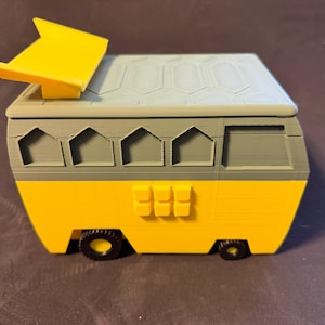 May include: A yellow and gray toy camper van with a yellow sunshade. The van has black wheels and a gray roof with a geometric pattern. The body of the van is yellow with gray accents and windows.