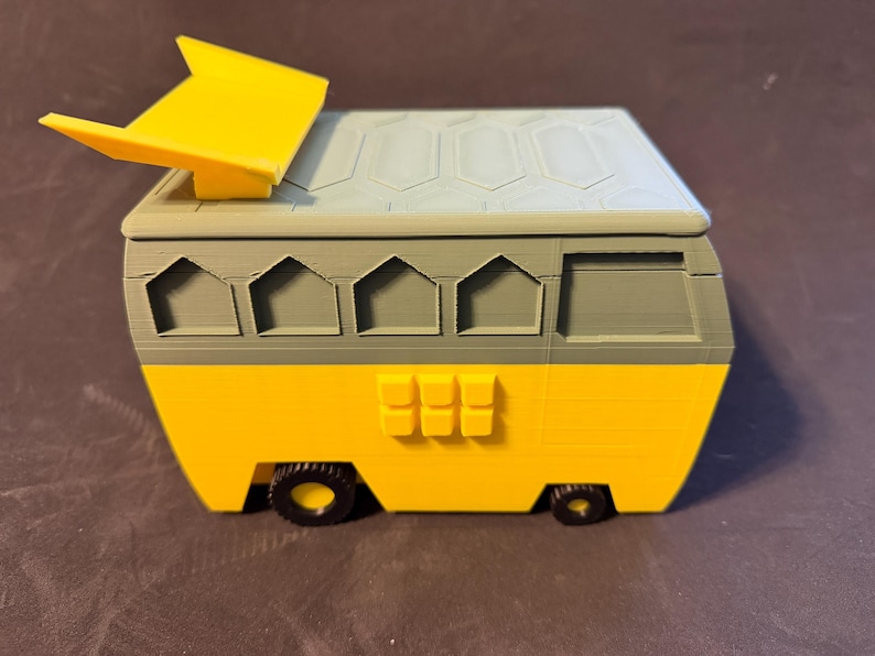 May include: A yellow and grey toy camper van with a yellow sunshade. The van has black wheels and a grey roof with a geometric pattern. The body of the van is yellow with grey accents and windows.
