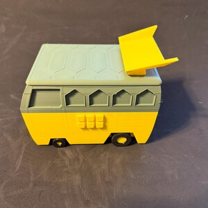 May include: A 3D-printed toy van in yellow, green, and light blue. The van has a rectangular body with a sloped roof and a yellow spoiler. The wheels are black, and the van has several windows and a door.