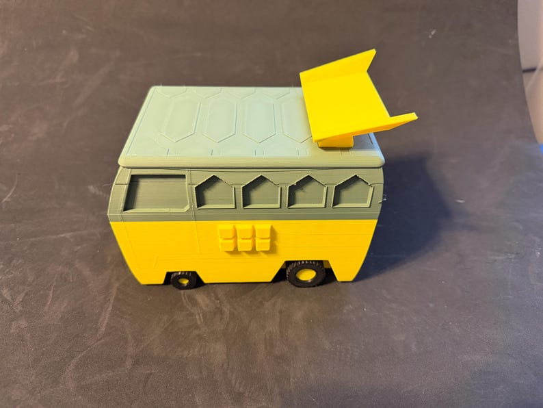 May include: A 3D-printed toy van in yellow, green, and light blue. The van has a rectangular body with a sloped roof and a yellow spoiler. The wheels are black, and the van has several windows and a door.