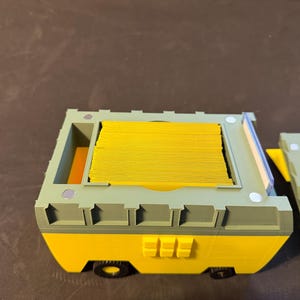 May include: A yellow and green 3D-printed storage container with a stack of yellow cards inside. The container has a removable lid and small black wheels. The design features a combination of rounded and angular shapes.