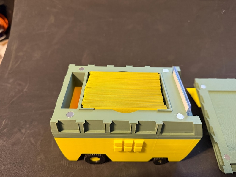 May include: A yellow and green 3D-printed storage container with a stack of yellow cards inside. The container has a removable lid and small black wheels. The design features a combination of rounded and angular shapes.