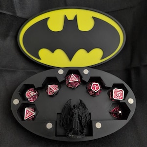 Könnte beinhalten: Eine offene, schwarze Würfelbox mit gelbem Batman-Logo, die rote und schwarze Würfel zeigt. Im Inneren befindet sich eine kleine Engelsfigur. Der Hintergrund ist dunkel.
