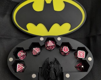 May include: An open black dice case with a yellow Batman logo on the lid. Inside, there are red and black dice. A small angel figurine is in the centre of the case. The background is dark.