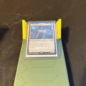 May include: A Magic: The Gathering card, "Giada, Font of Hope," is displayed in a protective sleeve, held by a yellow stand on a green base. The card features an illustration and text describing its abilities.