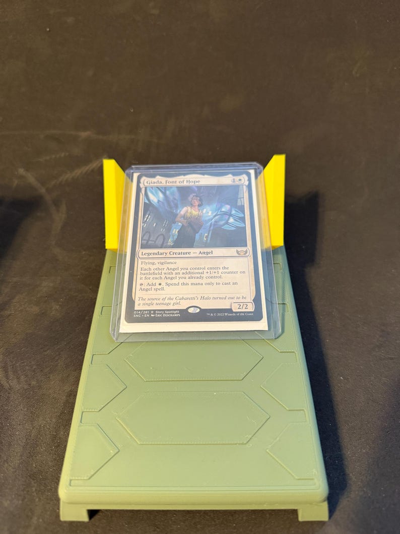 May include: A Magic: The Gathering card, "Giada, Font of Hope," is displayed in a protective sleeve, held by a yellow stand on a green base. The card features an illustration and text describing its abilities.