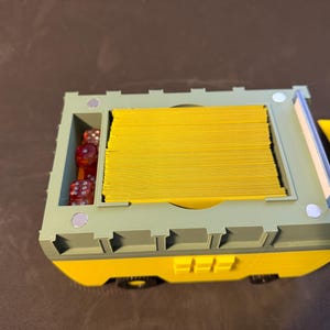 May include: A two-tone, yellow and green, game card and dice storage box. The box is rectangular with a compartment for dice and a larger compartment filled with yellow cards. The box has small wheels and a magnetic closure.
