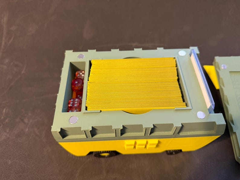 May include: A two-tone, yellow and green, game card and dice storage box. The box is rectangular with a compartment for dice and a larger compartment filled with yellow cards. The box has small wheels and a magnetic closure.