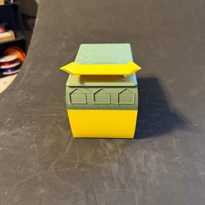 May include: A 3D-printed box in shades of yellow, green, and teal. The box has a rectangular shape with a yellow spoiler on top. The front of the box features three house-shaped cutouts. The box is sitting on a dark surface.