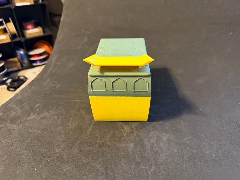 May include: A 3D-printed box in shades of yellow, green, and teal. The box has a rectangular shape with a yellow spoiler on top. The front of the box features three house-shaped cutouts. The box is sitting on a dark surface.