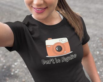 May include: Black t-shirt featuring a cartoon camera design in orange and white. The text "Don't be Negative" is printed below the camera. The shirt is made of a soft material.