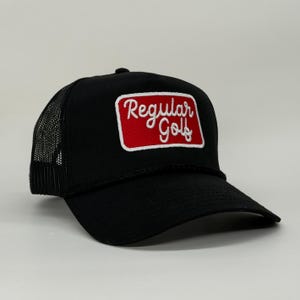 May include: Black trucker hat with a mesh back and a curved brim. The front features a rectangular patch with the words "Regular Golf" in white script on a red background.