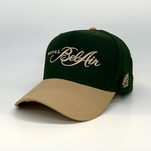 Może przedstawiać: Zielono-beżowa czapka z daszkiem z haftowanym na przodzie napisem "Hotel Bel Air" w kolorze złotym. Po prawej stronie czapki znajduje się małe haftowane złote listowie.