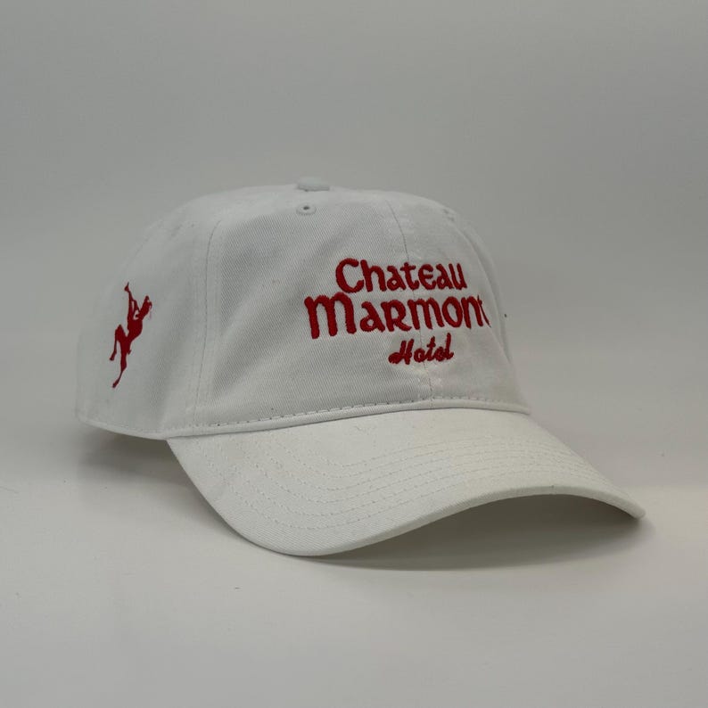 May include: White baseball cap with red embroidered text that reads "Chateau Marmont Hotel." A red graphic of a person on a horse is on the left side of the cap. The cap has a curved brim and a classic six-panel design.