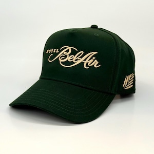 May include: A dark green baseball cap with the words "HOTEL Bel Air" embroidered in a cream-colored script across the front. A cream-colored leaf design is embroidered on the side.