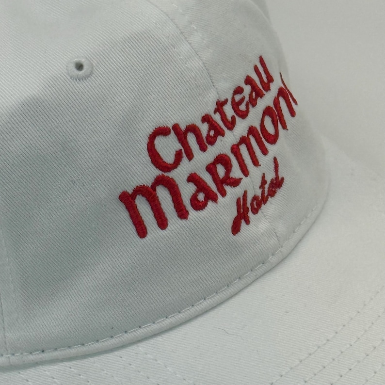May include: A white baseball cap with the red embroidered text "Chateau Marmont Hotel". The cap has a curved brim and a small ventilation hole on the top. The hat is made of a soft, durable fabric.