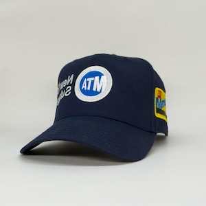 May include: A navy blue baseball cap with a curved brim. The cap features a circular patch with the letters "ATM" in white and blue. The text "New York" is also visible. A small patch with text is on the side.