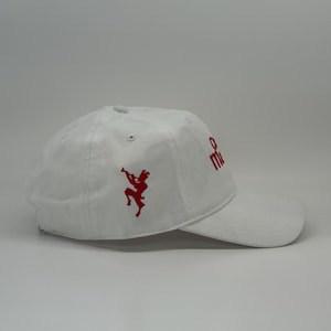 May include: White baseball cap with a curved brim. The cap features red embroidered designs, including a figure playing a horn and the letters "m" and "e". The cap is made of a soft, durable material.