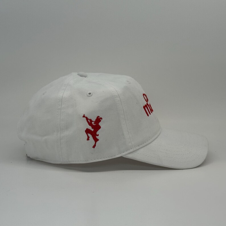 May include: White baseball cap with a curved brim. The cap features red embroidered designs, including a figure playing a horn and the letters "m" and "e". The cap is made of a soft, durable material.