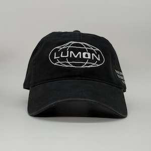 May include: Black baseball cap with the word "LUMON" in white, inside a white oval with a globe design. The cap has a curved brim and a slightly distressed appearance. The text "MACRO ERINE" is on the side.