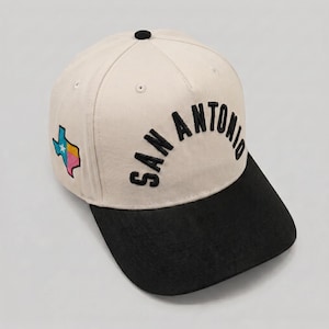 May include: A cream-colored baseball cap with a black brim and a black button on top. The front features the text SAN ANTONIO embroidered in black. The side displays an embroidered map of Texas in blue, white, and pink with a white star.