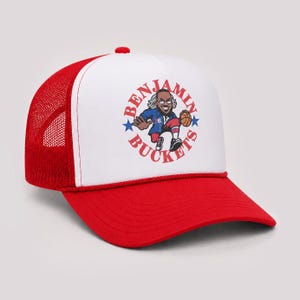 May include: A red and white trucker hat featuring a graphic of a basketball player in a blue jersey with the text BENJAMIN BUCKETS arranged in a circle around the figure, accented by two blue stars on the left and right sides of the design.