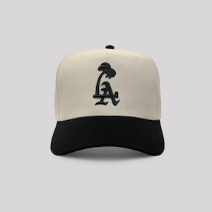Könnte beinhalten: Baseballcap mit cremefarbener Krone und schwarzem Schirm. Auf der Vorderseite der Kappe befindet sich ein schwarzes gesticktes Design der Buchstaben "LA" mit einer Palme. Die Kappe hat einen gebogenen Schirm und einen Knopf oben.
