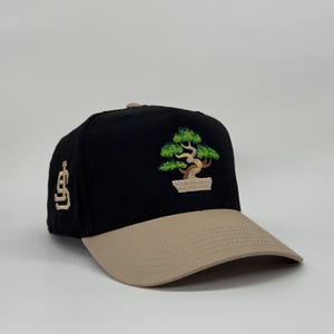 May include: Black baseball cap with a tan brim. The cap features an embroidered bonsai tree design in green, brown, and tan. The left side of the cap has a tan logo.