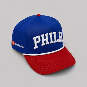 Puede incluir: Una gorra de béisbol azul con visera roja y un detalle de cuerda blanca. La parte frontal presenta la palabra PHILA en letras blancas bordadas. El lateral muestra un cuadrado naranja con una B blanca y el texto NRG Station en blanco.