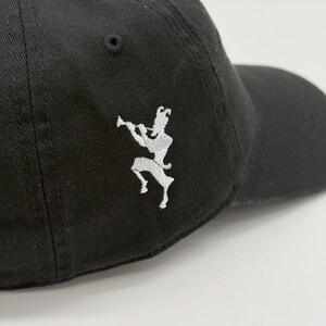 May include: Black baseball cap with a curved brim. The cap features a white embroidered design of a figure playing a wind instrument on the side. The cap is made of a durable fabric and has a classic, casual style.