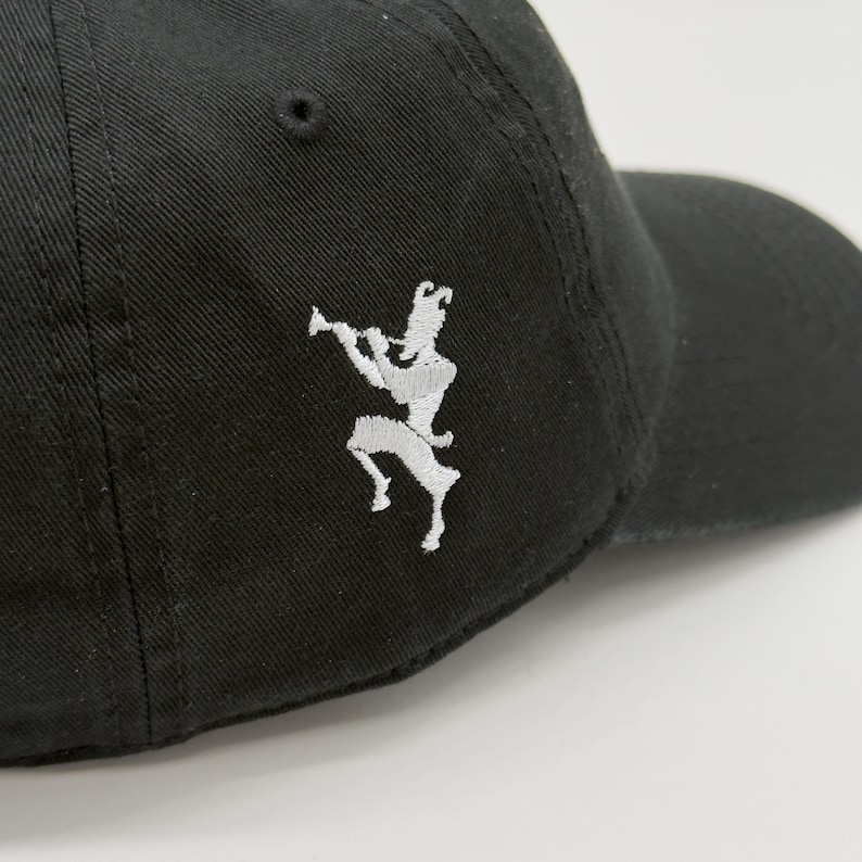 May include: Black baseball cap with a curved brim. The cap features a white embroidered design of a figure playing a wind instrument on the side. The cap is made of a durable fabric and has a classic, casual style.