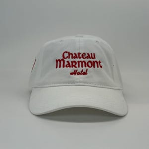 May include: White baseball cap with the embroidered text "Chateau Marmont Hotel" in red. The cap has a curved brim and a classic six-panel design, suitable for casual wear.