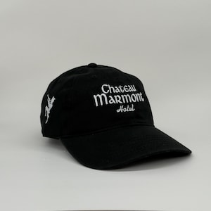 May include: Black baseball cap with the text "Chateau Marmont Hotel" embroidered in white. The cap also features a white embroidered design on the side. The hat has a curved brim and an adjustable strap.