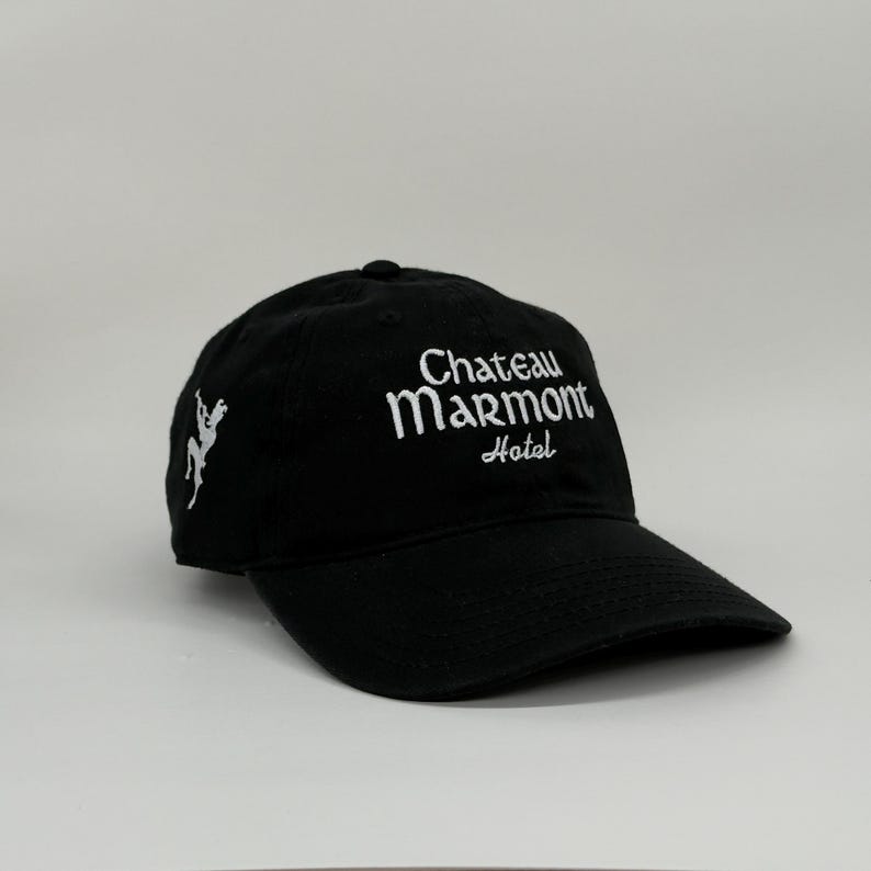 May include: Black baseball cap with the text "Chateau Marmont Hotel" embroidered in white. The cap also features a white embroidered design on the side. The hat has a curved brim and an adjustable strap.
