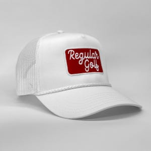 Pode incluir: Boné de caminhoneiro branco com um patch retangular vermelho bordado com as palavras "Regular Golf" em fonte cursiva. O boné apresenta uma parte traseira de malha branca, uma aba curva e um detalhe de corda branca.