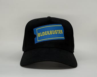Op de afbeelding: Zwarte baseballpet met een rechthoekige patch met het woord "BLOCKBUSTER" in gele letters op een blauwe achtergrond. De patch heeft een gele rand en een kleine inkeping aan de linkerkant.