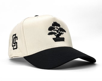 Pode incluir: Um boné de beisebol com coroa creme e aba preta. O boné apresenta um design bordado em preto de uma árvore bonsai na frente e as letras "CS" na lateral. O chapéu está sobre um fundo branco.