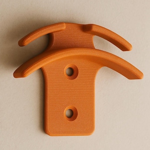May include: An orange wall mount with a distinctive shape. It has two curved arms at the top and a rectangular base with two circular holes. The mount is made of a textured material, and is a shade of orange.