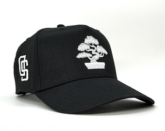 Pode incluir: Boné de beisebol preto com um design de bonsai bordado em branco na frente. O boné também apresenta um logotipo branco bordado na lateral. Possui uma aba curva e um ajuste estruturado.