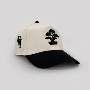 May include: Cream-colored baseball cap with a black brim. The cap features a black embroidered bonsai tree design on the front and the letters "VT" on the side. The cap has a curved brim and a button on top.