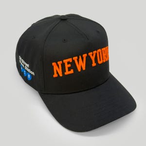 Pode incluir: Um boné de beisebol preto com o texto NEW YORK bordado em laranja brilhante na frente. A lateral do boné exibe o texto 34 Street Penn Station com ícones circulares azuis contendo as letras A, C e E em branco.