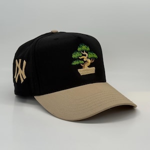 Könnte beinhalten: Eine schwarze und beige Baseballkappe mit einem grünen Bonsai-Baum, der auf der Vorderseite gestickt ist. Die Kappe hat ein golden gesticktes "NY" an der Seite.