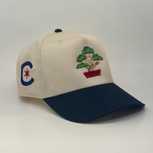 May include: A beige baseball cap with a navy blue brim. The front of the cap features an embroidered 'C' with a red star and a green bonsai tree in a red pot.
