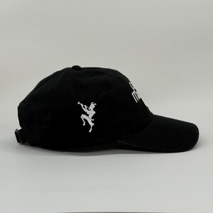 May include: Black baseball cap with a curved brim and adjustable strap. The cap features a white embroidered design of a figure playing a wind instrument on the side and white text on the front.