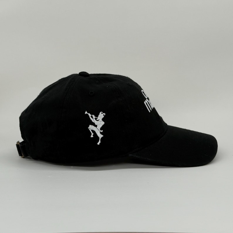May include: Black baseball cap with a curved brim and adjustable strap. The cap features a white embroidered design of a figure playing a wind instrument on the side and white text on the front.